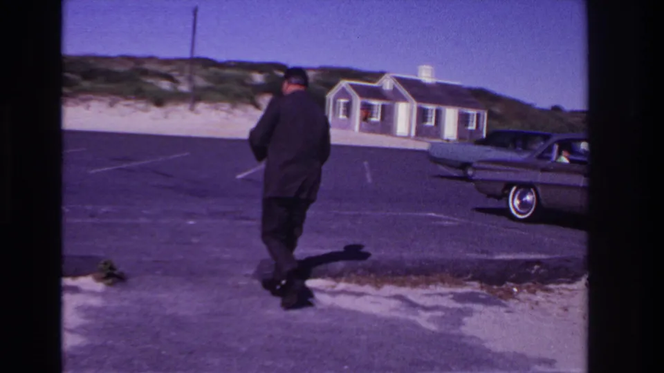 A man in a dark suit continues across the Rockaway Beach parking lot, with a parked mid-century car and dune-backed buildings in view.