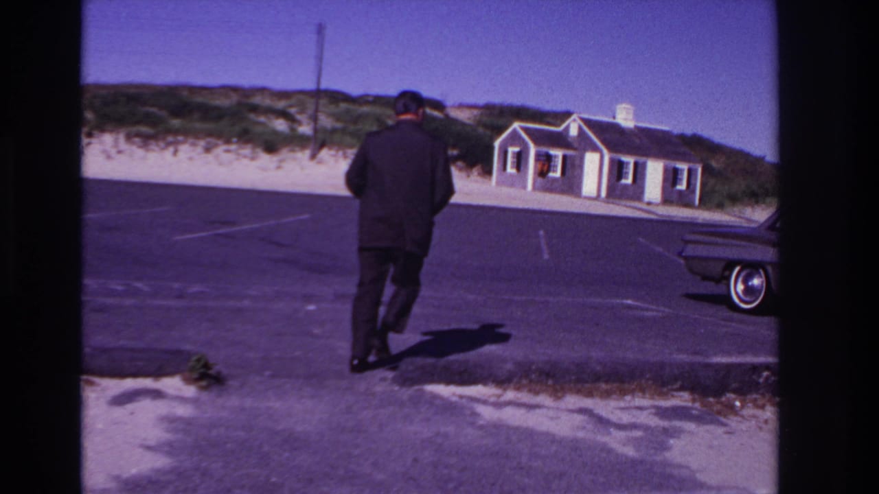 In Rockaway Beach, New York, 1967, a suited man walks across an asphalt beach lot with dunes, a small building, and part of a classic car visible.