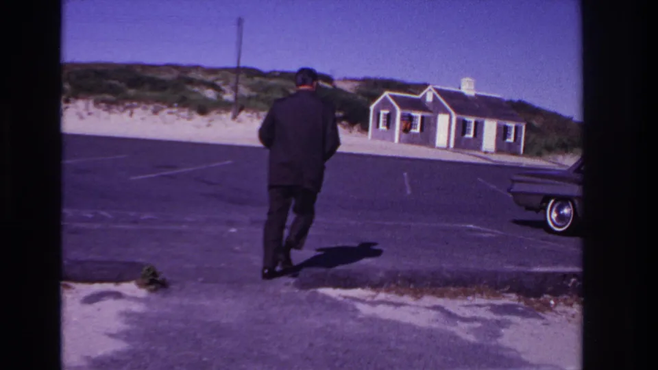In Rockaway Beach, New York, 1967, a suited man walks across an asphalt beach lot with dunes, a small building, and part of a classic car visible.