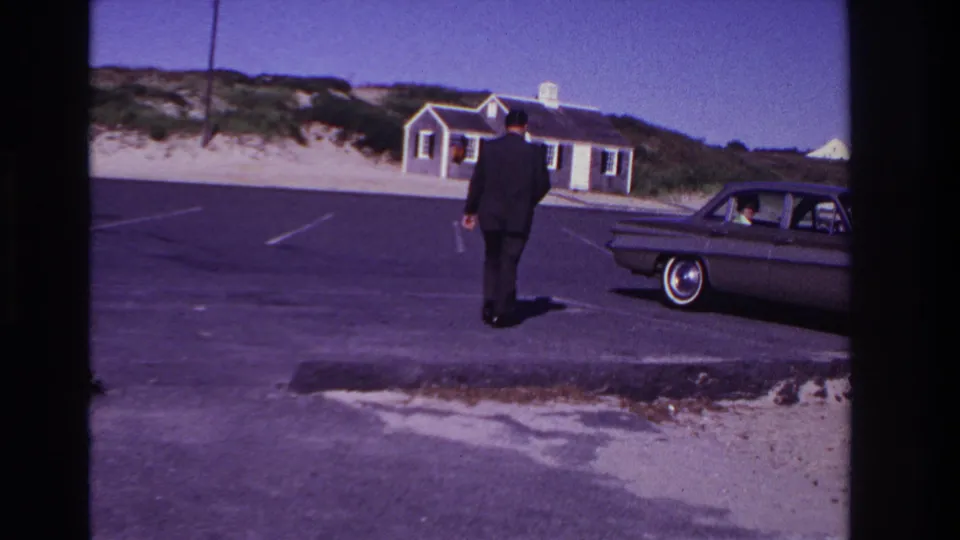 A suited man approaches a classic car in the Rockaway Beach lot while a passenger is visible inside, with beach dunes and a small building behind.