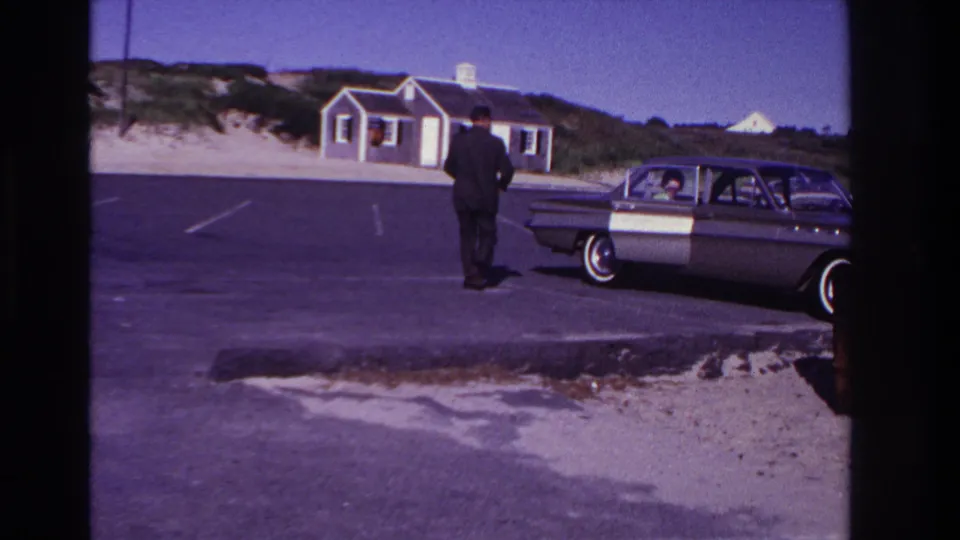 At Rockaway Beach in 1967, the suited man stands near the side of the classic car, with a passenger, dunes, and shore buildings visible.