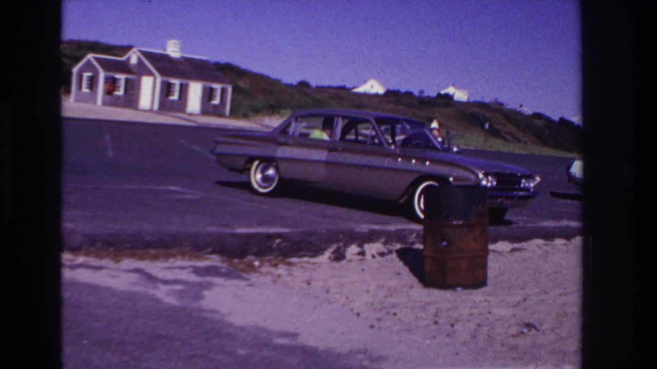 Classic sedan parked beside sand and pavement with a person visible near the right side of the car.