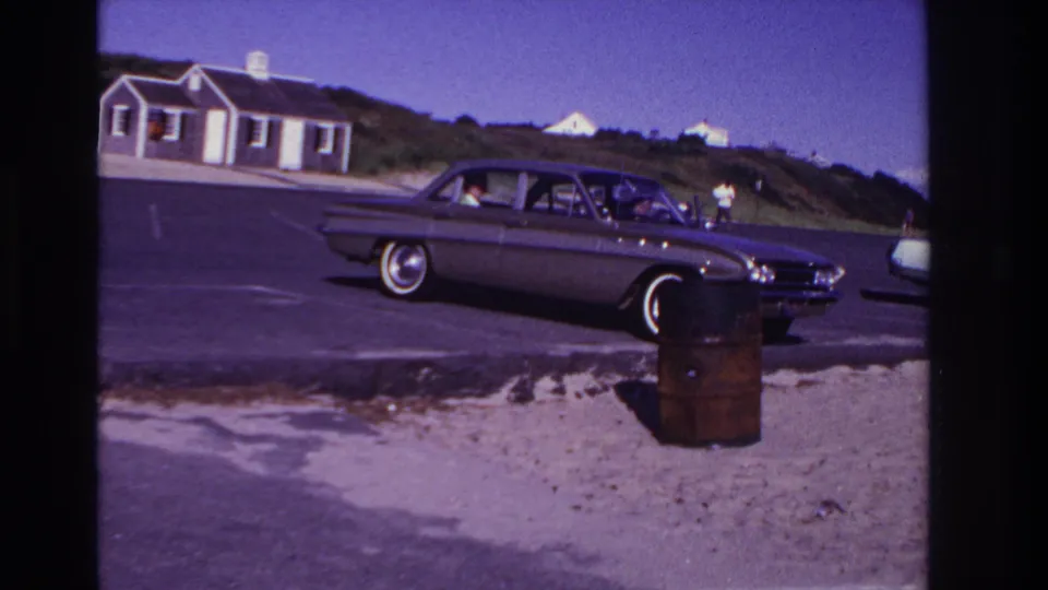 Rockaway Beach home-movie frame showing the sedan in profile as a person stands near it in the coastal parking lot.