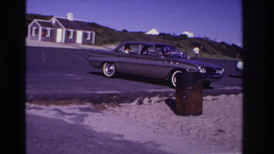 Rockaway Beach home-movie frame from 1967 showing a classic sedan near a sandy roadside, dunes, and a small seaside building.
