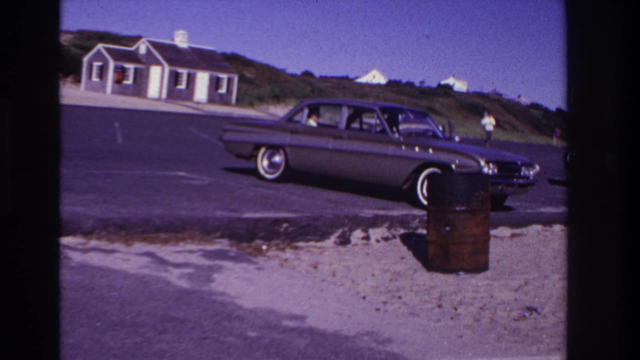 Classic sedan on a beach road with dunes, small buildings, and a person standing in the background.