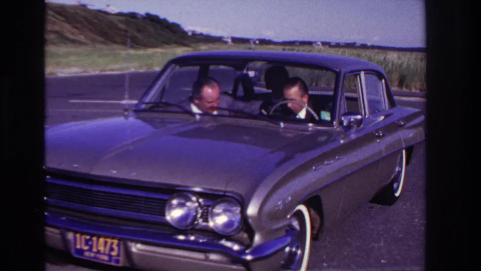 Close 1967 Rockaway Beach home-movie view of two adult men in business attire inside a classic sedan near the coastal road.