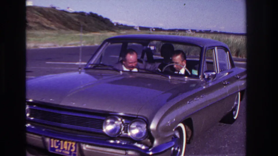 Front view of a classic sedan at Rockaway Beach in 1967, with two adult men visible inside and dune grass beyond the road.