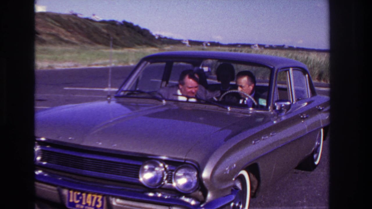 At Rockaway Beach in 1967, two adult men sit in a stopped dark 1960s sedan, looking downward through the windshield with dunes and sky beyond.