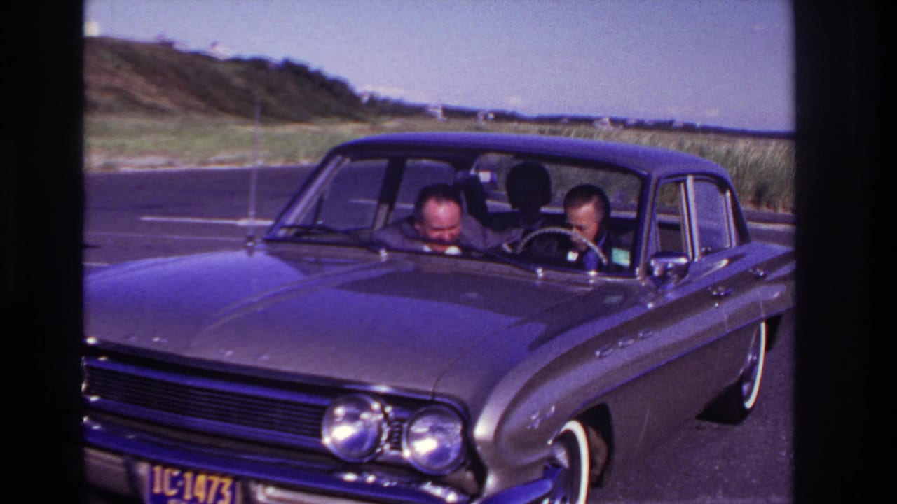 Two men lean forward inside a stopped sedan near dunes and open sky.