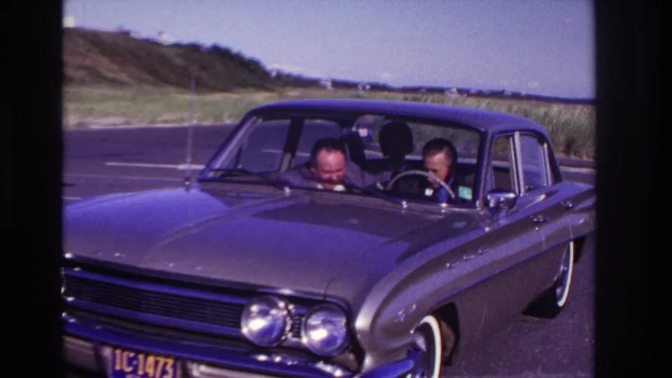 The driver and passenger lean forward in a dark 1960s sedan at Rockaway Beach, with the coastal road, dunes, and horizon visible behind them.