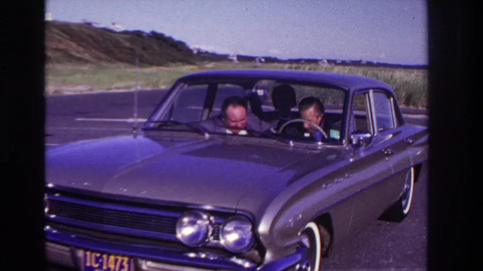 A 1967 Rockaway Beach home-movie frame shows two men in the front seat of a dark sedan, looking down as dunes and beach structures sit in the distance.