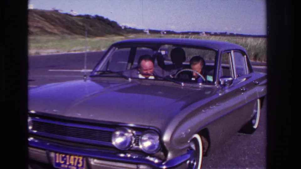 Two adult men sit low in a dark 1960s sedan at Rockaway Beach, still focused downward inside the car as the coastal landscape remains behind them.