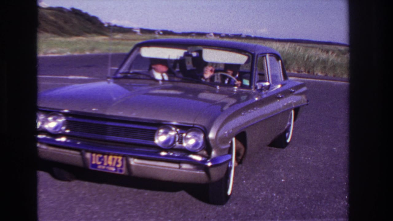 At Rockaway Beach in 1967, a dark classic sedan approaches the camera on a paved road, with the adult driver visible behind the wheel.