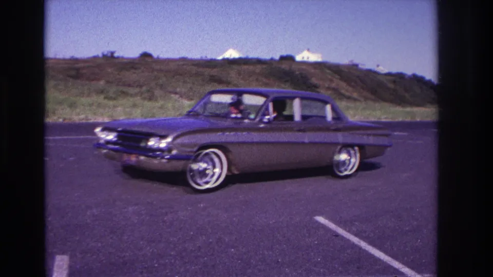 A dark mid-1960s sedan passes in side view at Rockaway Beach, with whitewall tires, chrome trim, and low houses in the background.