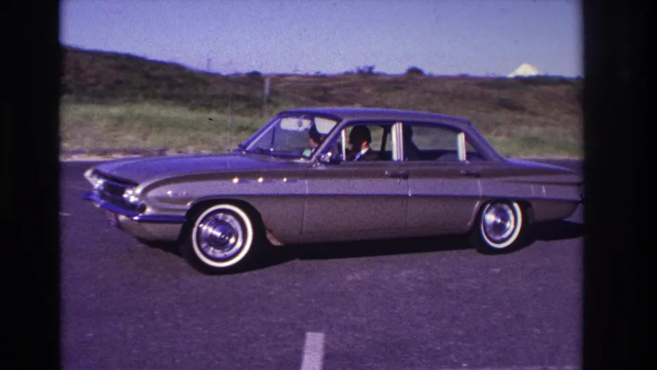 The sedan is shown in side profile on the Rockaway Beach road, with occupants visible through the windows and grass-covered terrain behind.