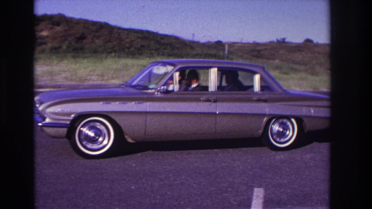 Light-colored 1960s sedan passing left to right in an empty parking lot.