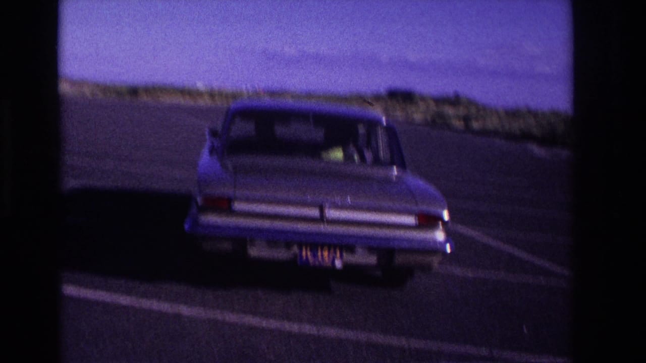 The sedan turns away from the camera in the Rockaway Beach lot, seen from behind with dunes and sky beyond the asphalt.