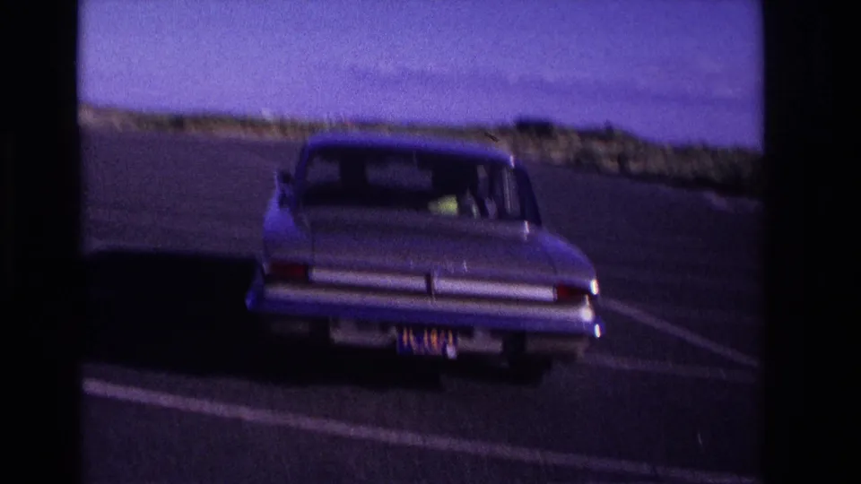 The sedan turns away from the camera in the Rockaway Beach lot, seen from behind with dunes and sky beyond the asphalt.