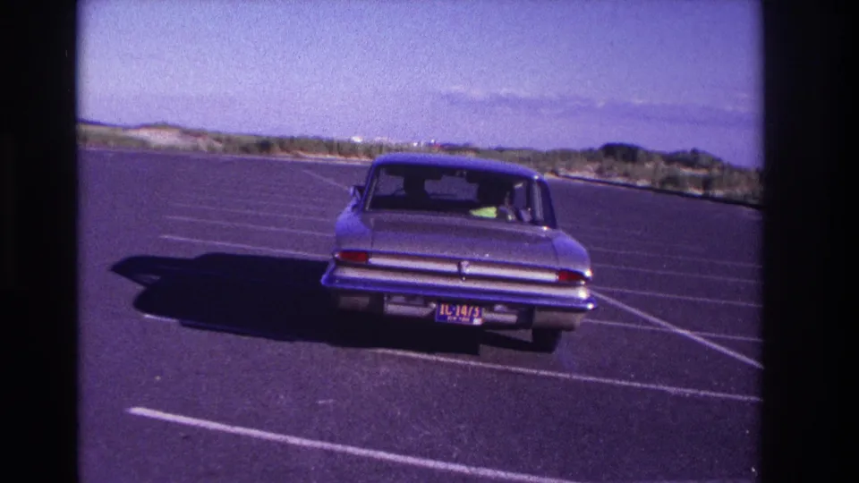 A rear view shows the light-colored sedan moving deeper into the open Rockaway Beach parking area, with painted lines crossing the asphalt.