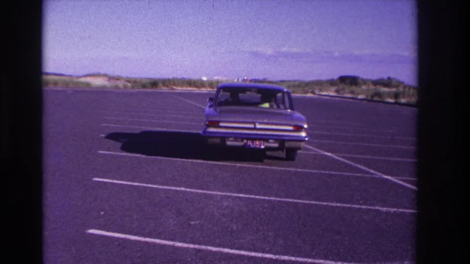 The sedan appears farther from the camera in the Rockaway Beach parking lot, surrounded by empty spaces, dunes, and open sky.