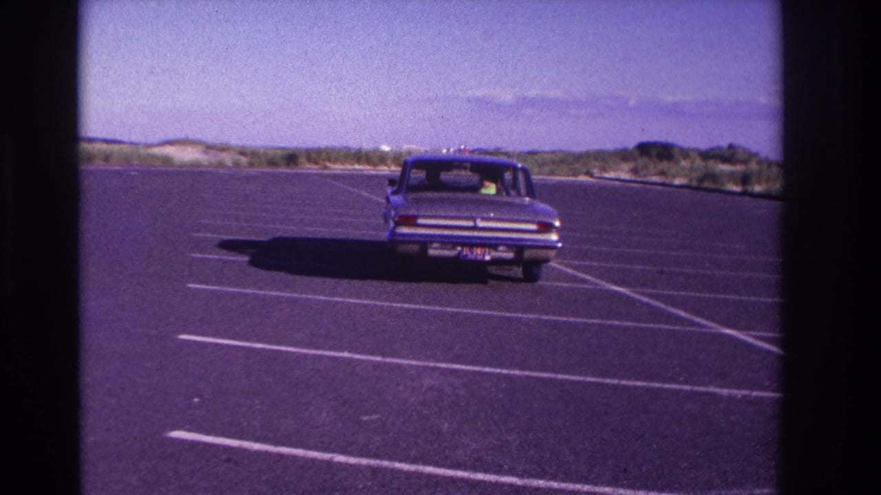 1967 Rockaway Beach home-movie frame showing a blue sedan moving away through a mostly empty beach parking lot.