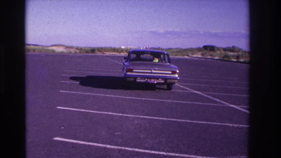 1967 Rockaway Beach home-movie frame showing a blue sedan moving away through a mostly empty beach parking lot.