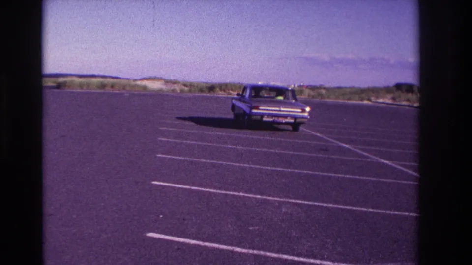 A rear view of the mid-1960s sedan receding across the striped lot, with beach dunes and sky in the background.