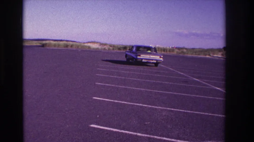 By the final frame, the sedan is distant in the Rockaway Beach lot, surrounded by open asphalt and low beach vegetation.