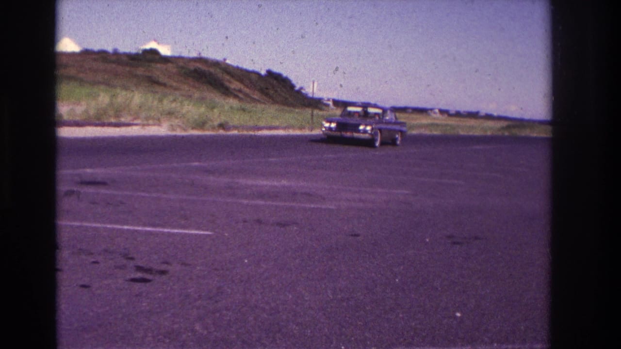 Dark sedan approaching across an empty asphalt parking lot with dunes and pale buildings behind it.