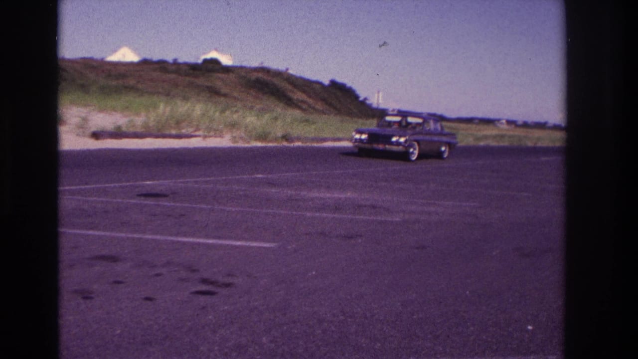 Home-movie view of a dark sedan moving across a mostly empty Rockaway Beach parking lot, with dunes and small buildings beyond.