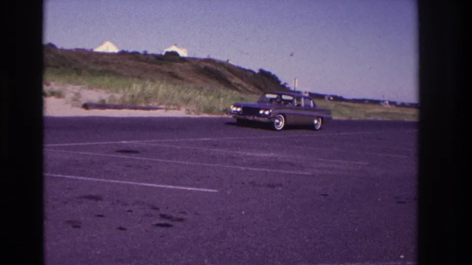 A dark mid-1960s sedan passes nearer the camera in a marked parking lot at Rockaway Beach, with dune grass behind it.