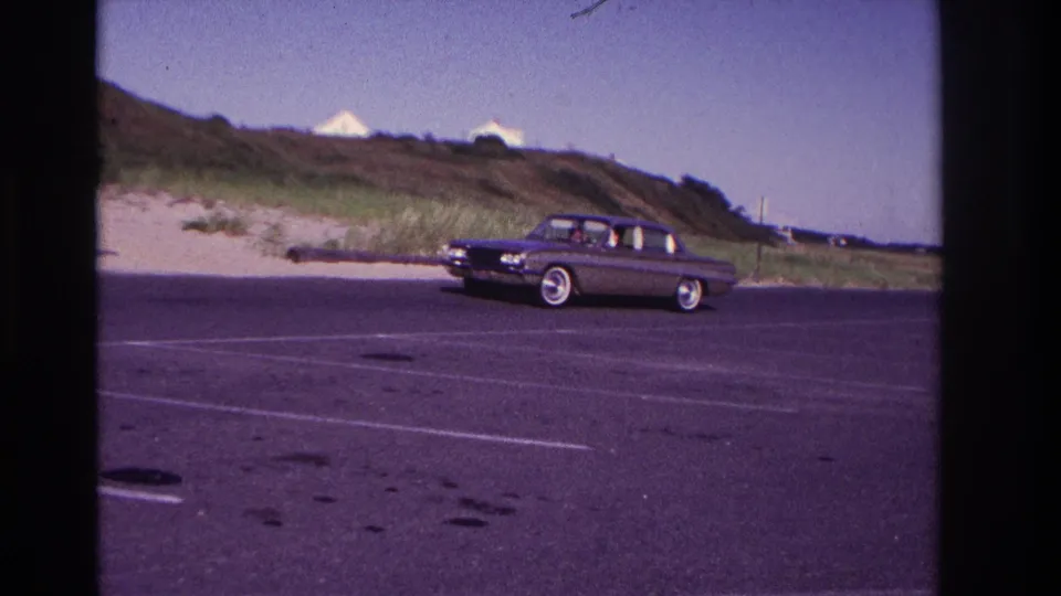 Side view of the dark sedan in the Rockaway Beach parking lot, with possible occupants visible through the windows but no clear faces.