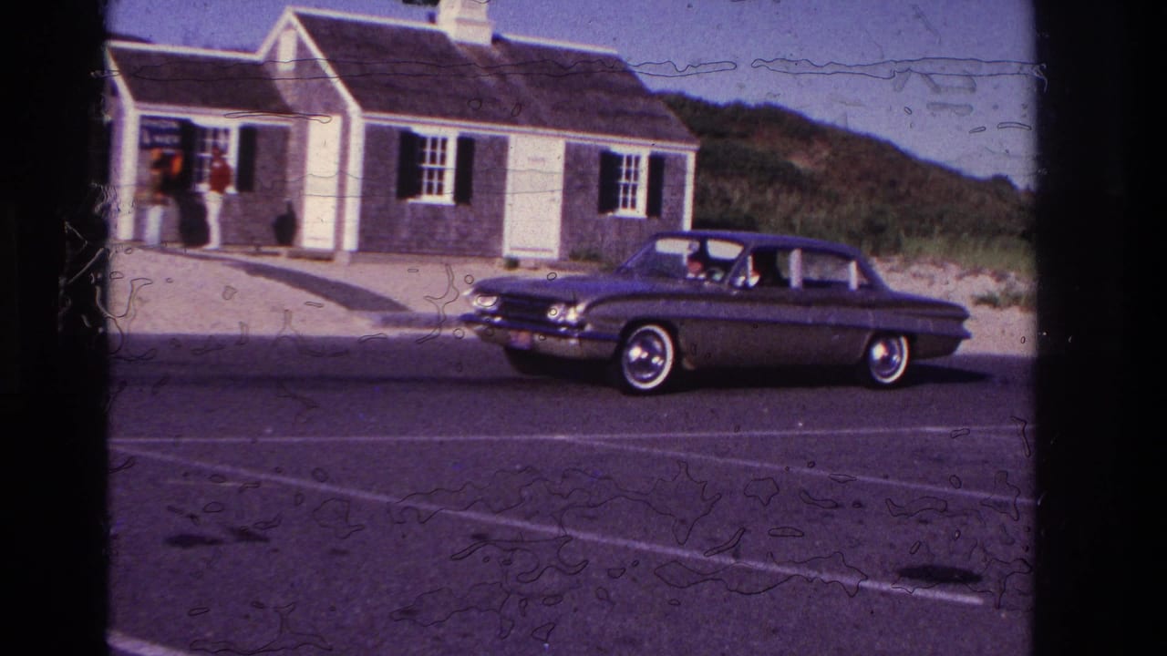 1967 Rockaway Beach home-movie frame showing a dark classic sedan passing a small shingled house with white trim.