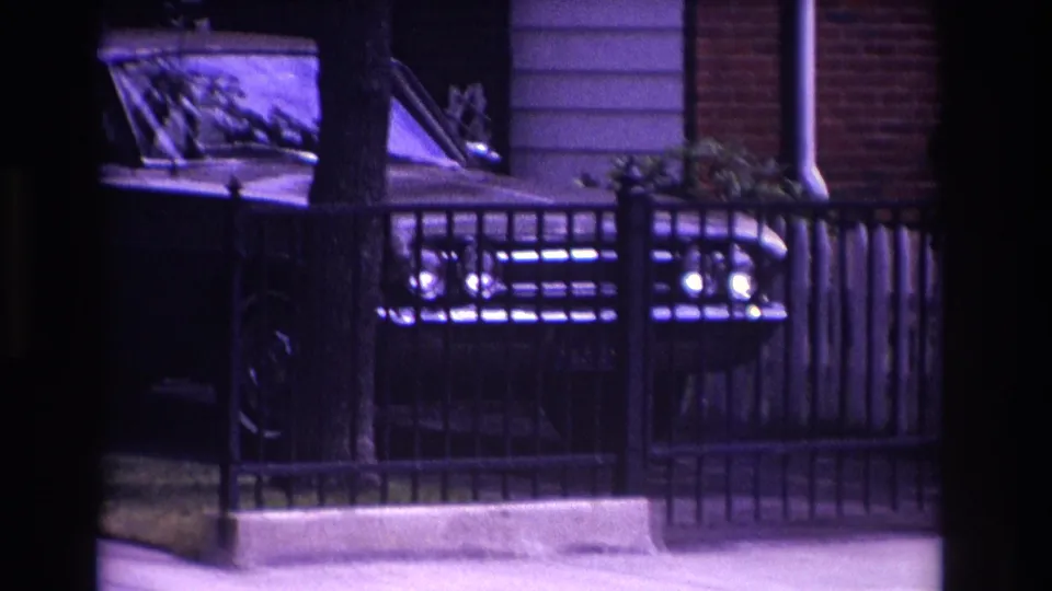 A close nighttime view shows the classic sedan behind a fence beside house siding and brickwork in the 1967 Rockaway Beach footage.
