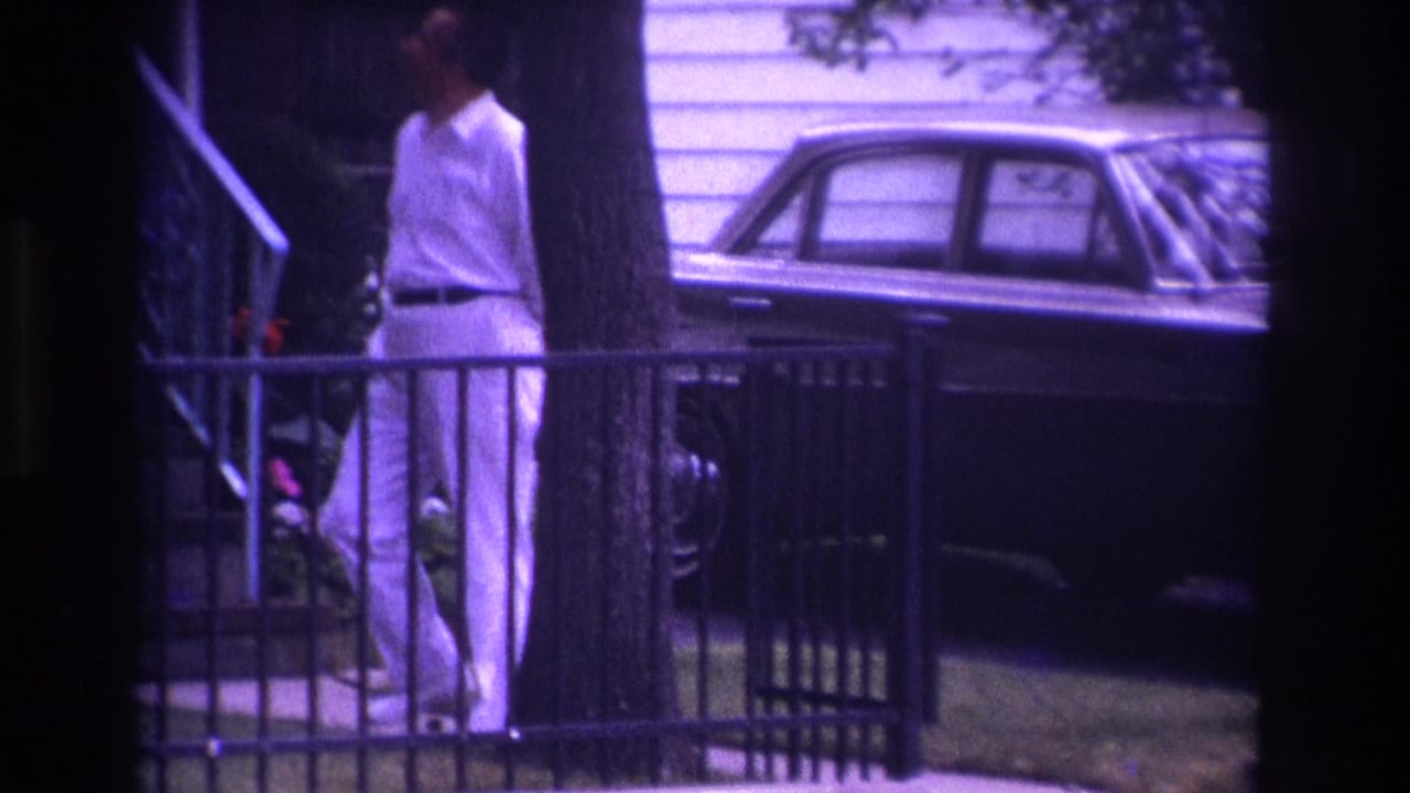 Man in a white shirt and trousers stands beside a railing with a sedan and house behind him.