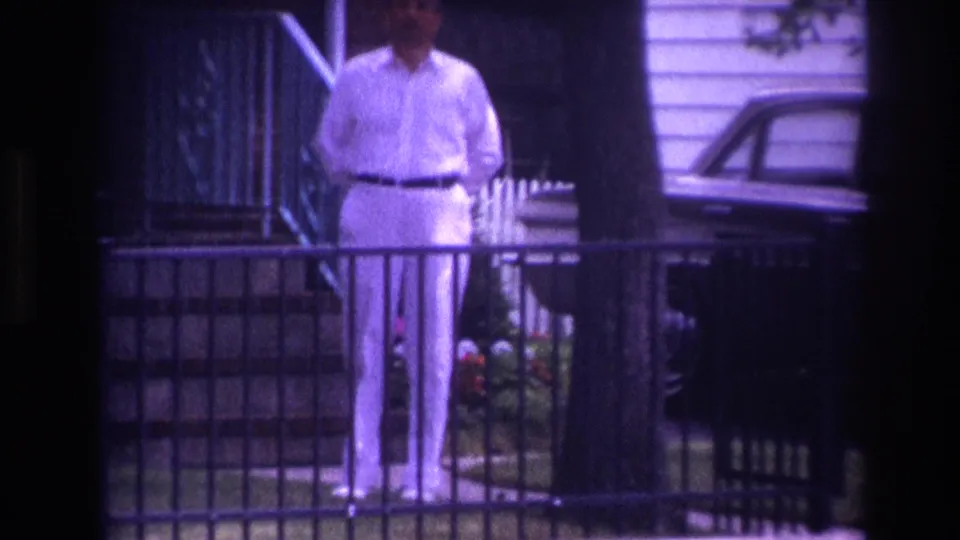 1967 Rockaway Beach home-movie frame showing a man in white behind a railing, with porch steps, flowers, a tree, and part of a parked car visible.