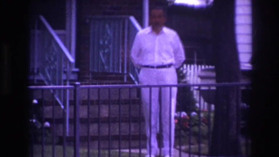 1967 Rockaway Beach frame with a man in white clothing standing behind a front railing, near porch steps, flowers, trees, and residential siding.