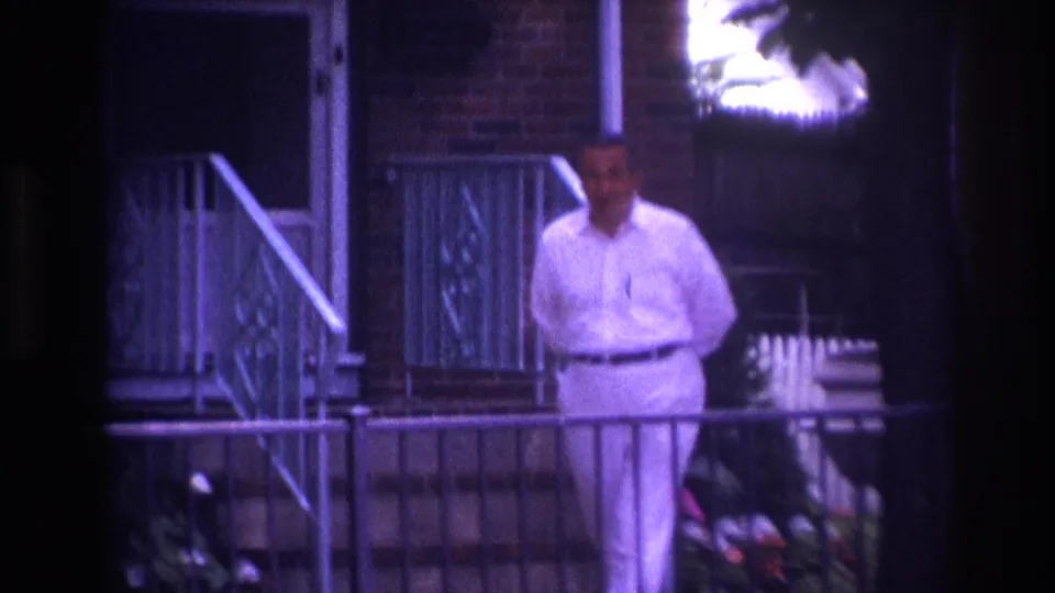 1967 Rockaway Beach home-movie frame showing a man in white clothing on a porch beside metal railings and a brick house exterior.