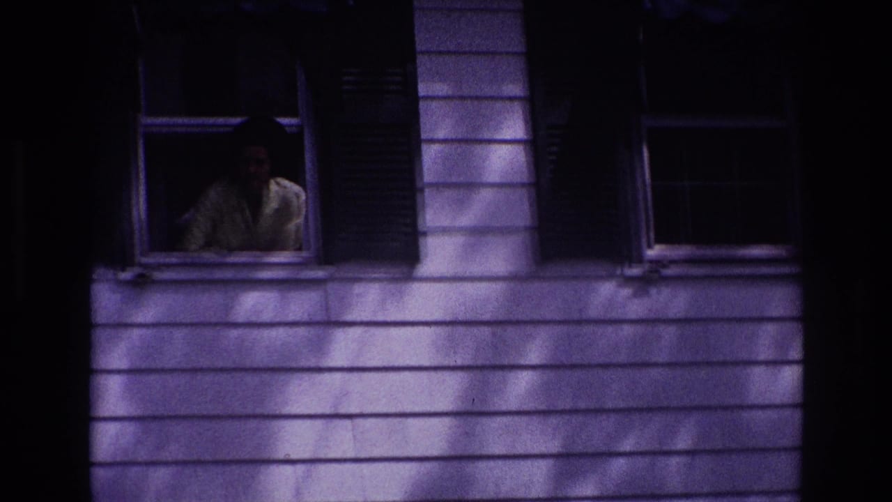 1967 Rockaway Beach frame with a woman partially visible in an upstairs window of a shadowed house exterior.