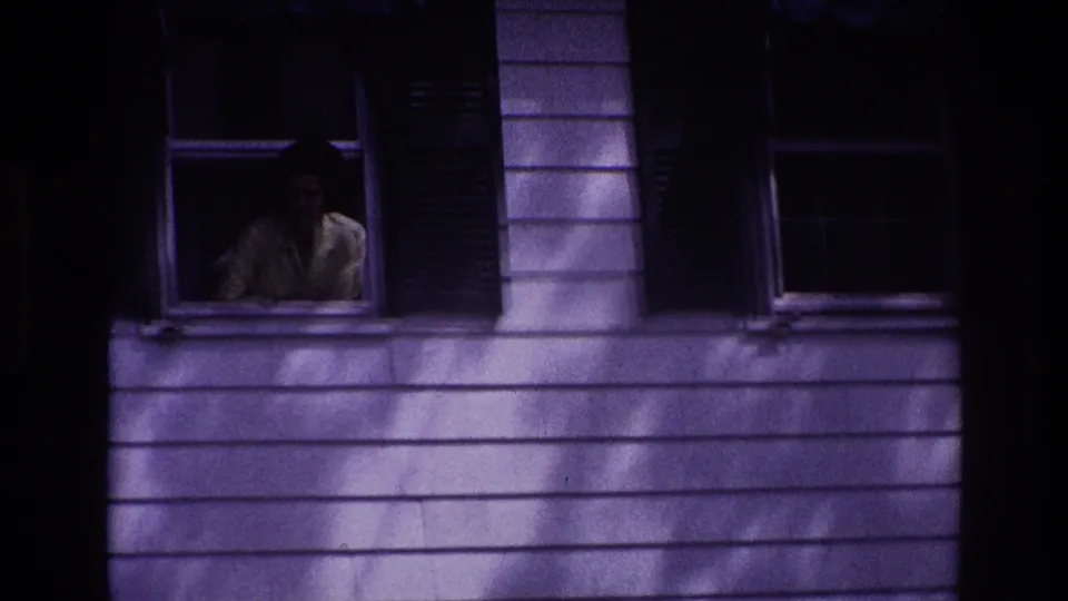 1967 Rockaway Beach frame with a woman partially visible in an upstairs window of a shadowed house exterior.