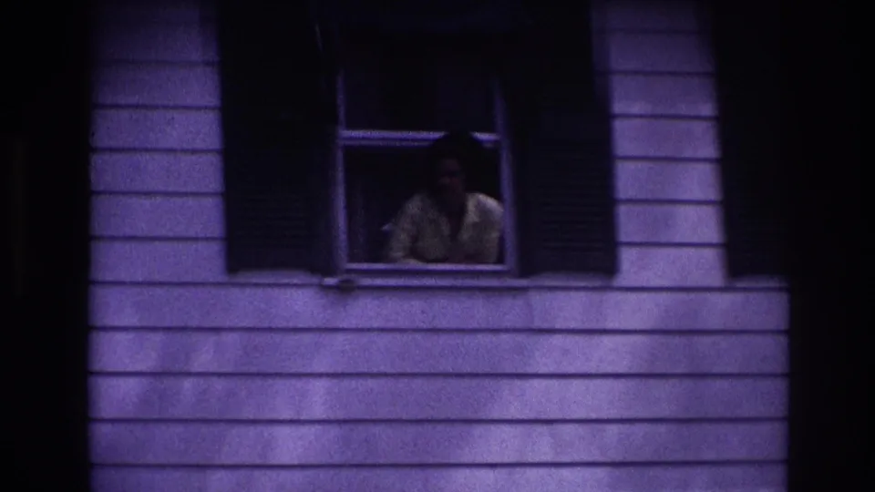 Vintage 8mm frame from Rockaway Beach showing a woman at a shuttered window on a house facade.