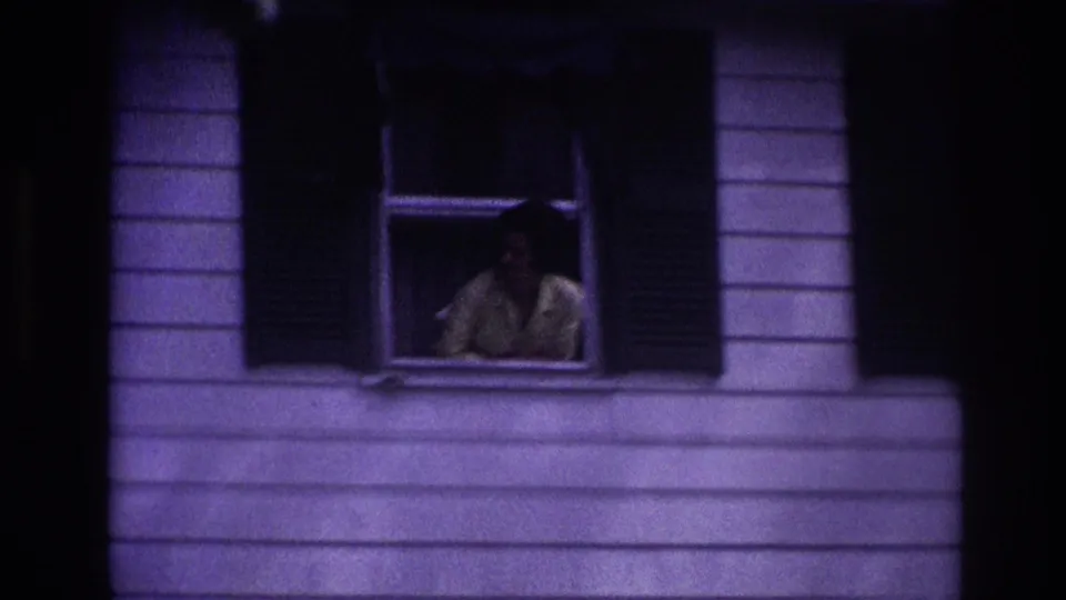 1967 Rockaway Beach 8mm frame showing the woman still at the window, surrounded by shutters, siding, and deep shadow.