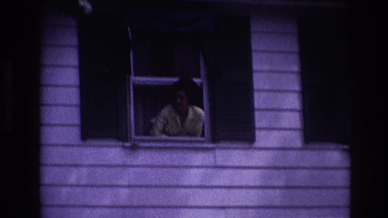 1967 Rockaway Beach home-movie frame showing a woman in a yellow shirt at an open window of a white-sided house.