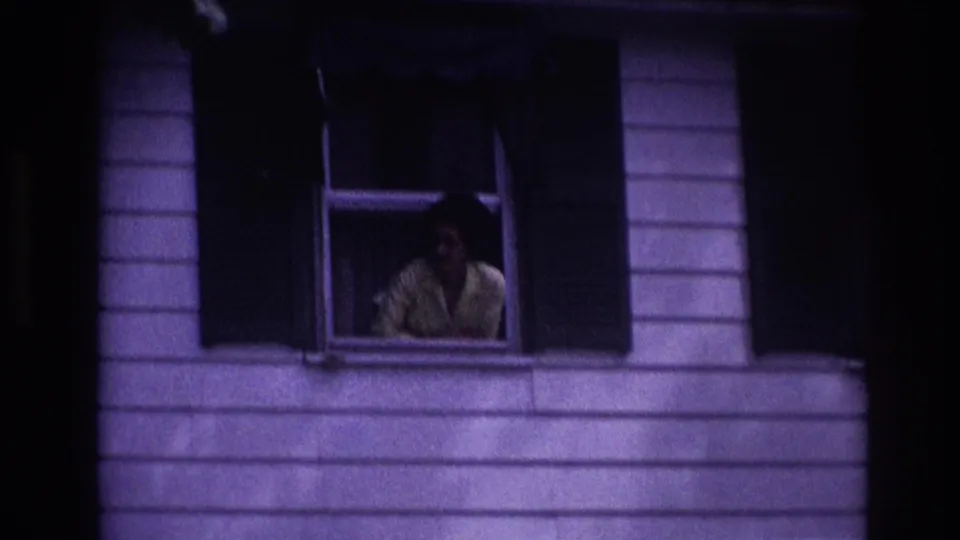 1967 Rockaway Beach home-movie frame showing a woman in a yellow shirt at an open window of a white-sided house.