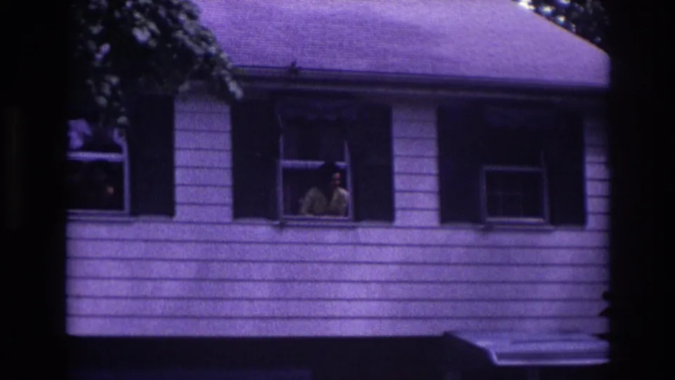 Wider 1967 Rockaway Beach frame showing a white-sided house exterior with the woman visible at one open window.