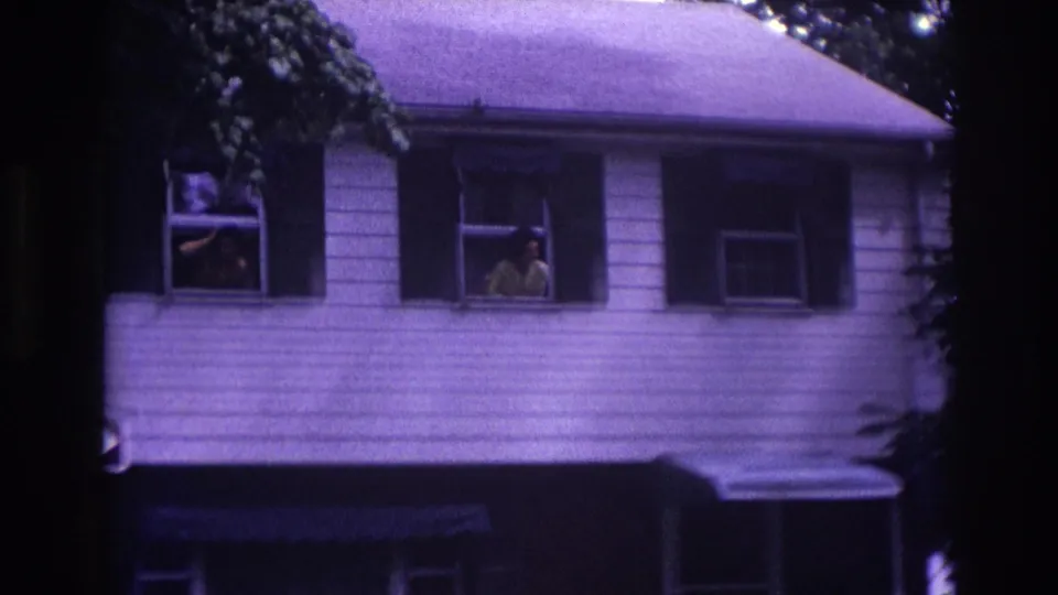 1967 Rockaway Beach home-movie frame showing two adults at separate second-story windows of a white-sided house.