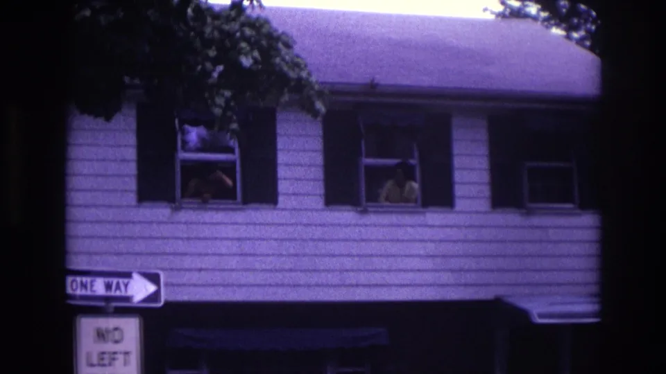 1967 home-movie frame of a white-sided house with people at upstairs windows and ONE WAY and NO LEFT signs below.