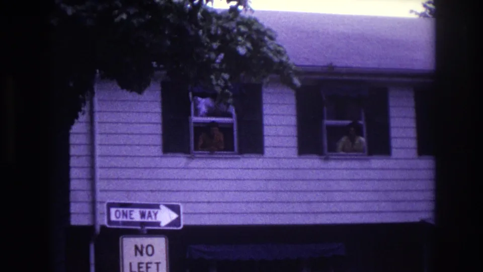 Rockaway Beach home-movie frame showing two adults in separate upstairs windows above ONE WAY and NO LEFT street signs.