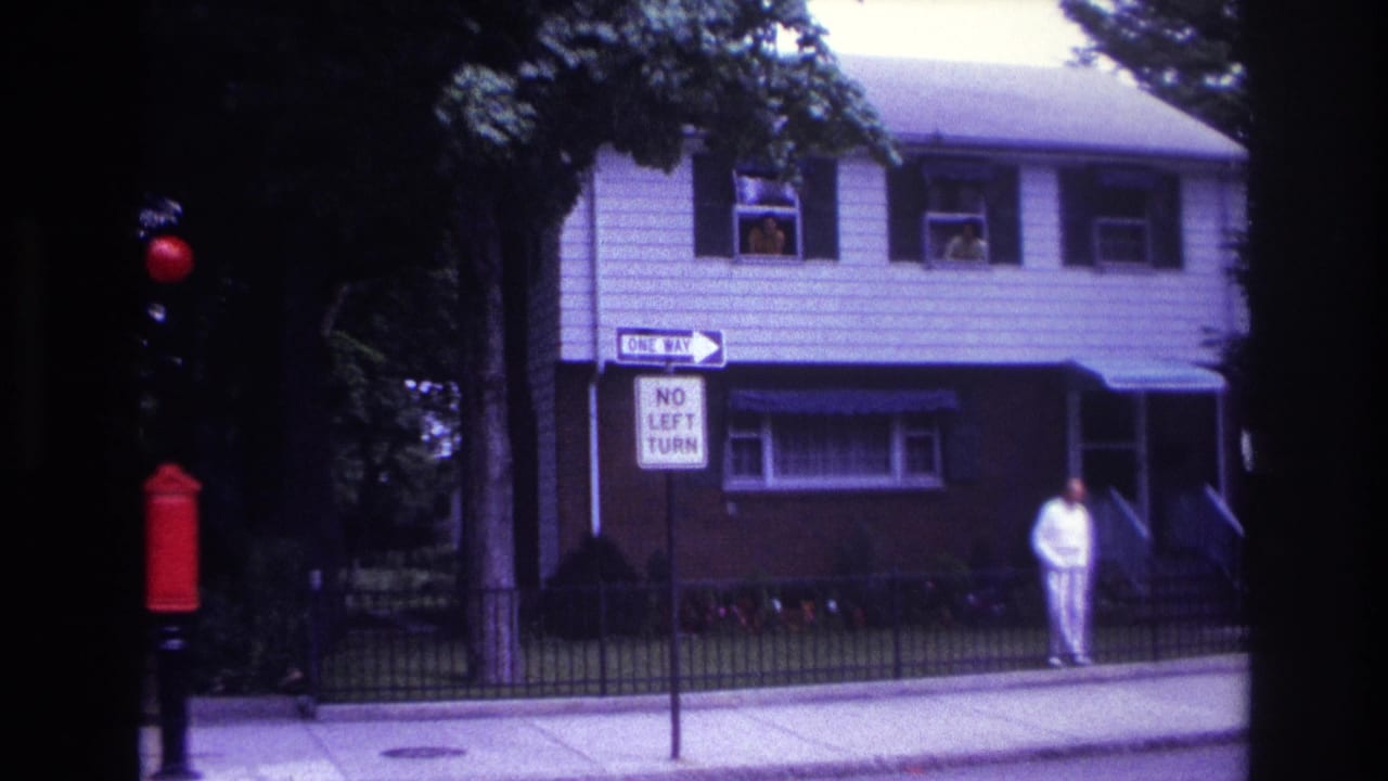 Man in white clothing standing near a corner house, street signs, fence, and red hydrant.