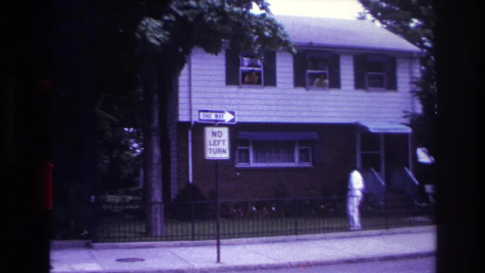 Home-movie frame from 1967 Rockaway Beach showing people in upper windows while a man in white stands outside the house.